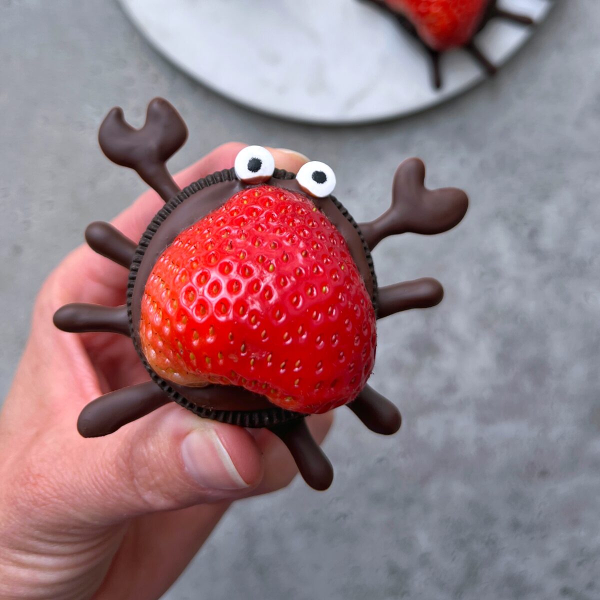 A hand holds a creative dessert bite made of a strawberry, Oreo, and chocolate details, decorated to look like a crab with candy eyes.