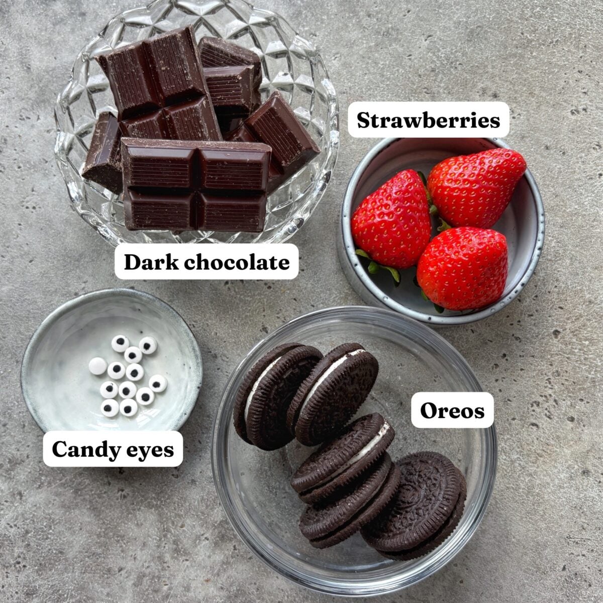 Four bowls with labeled ingredients including dark chocolate squares, whole strawberries, candy eyes, and Oreo cookies.