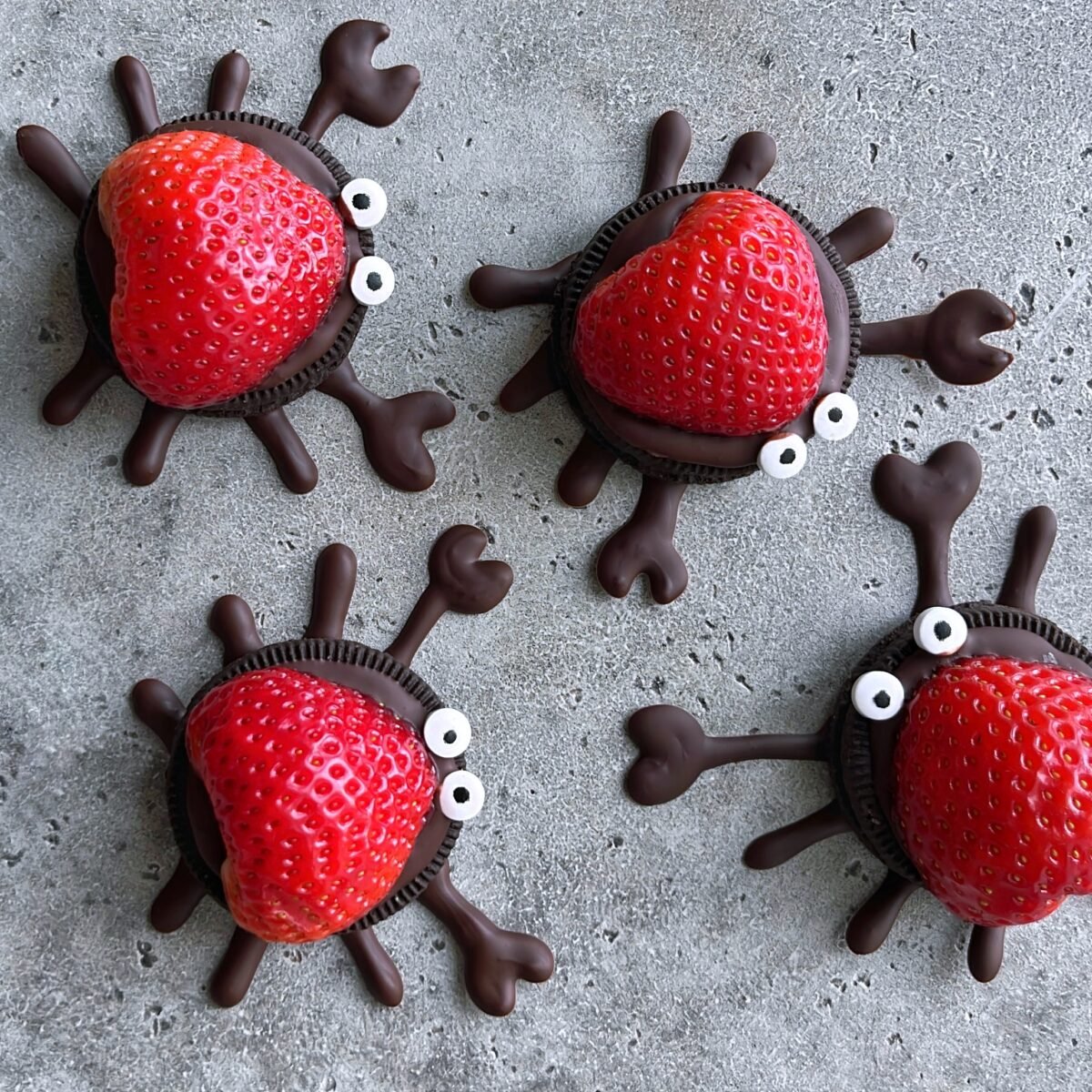 Four Oreo cookies topped with half strawberries, decorated with chocolate legs and candy eyes to resemble crabs, displayed on a gray textured surface.
