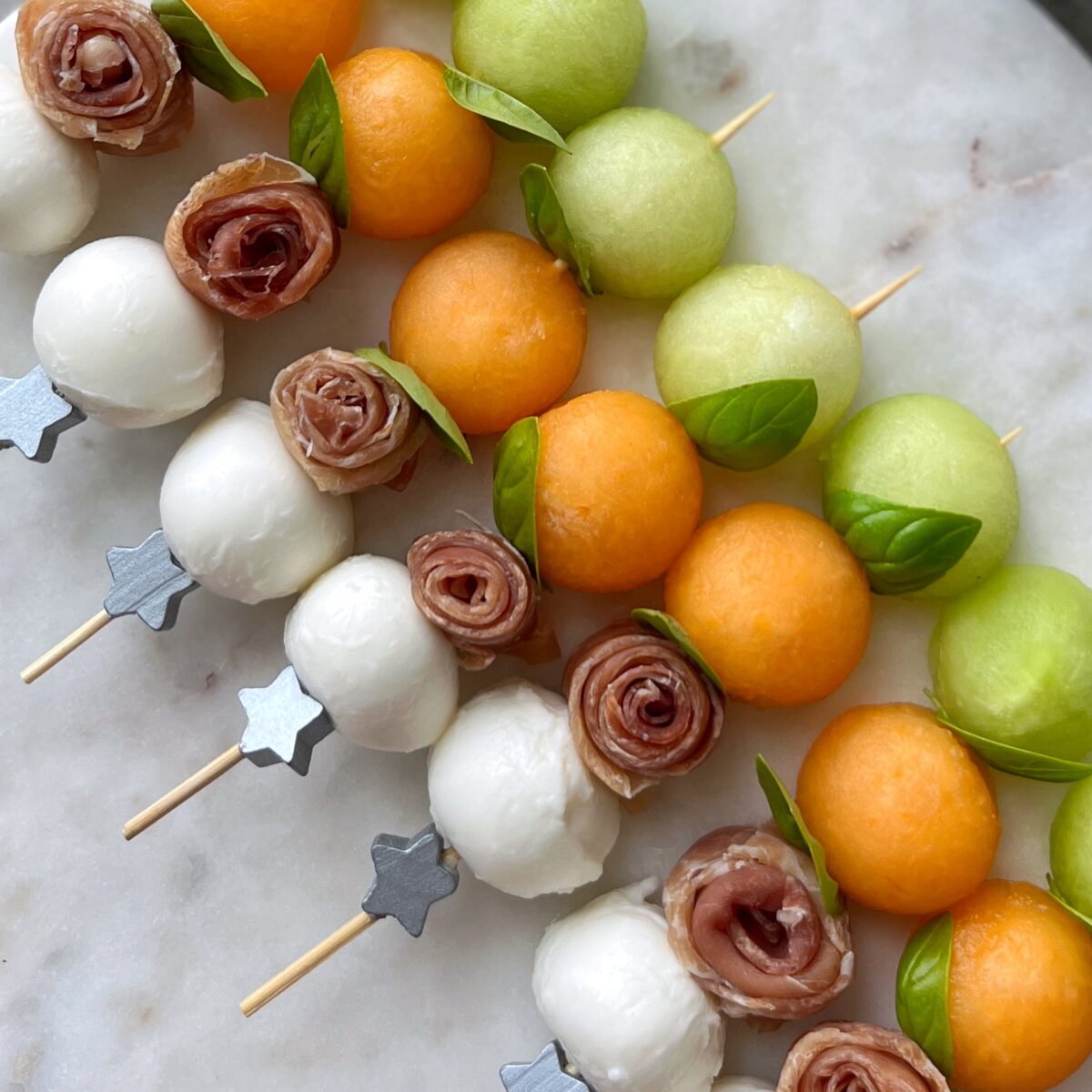 Skewers with mozzarella balls, cantaloupe, honeydew melon balls, prosciutto roses, and basil arranged on a marble surface.