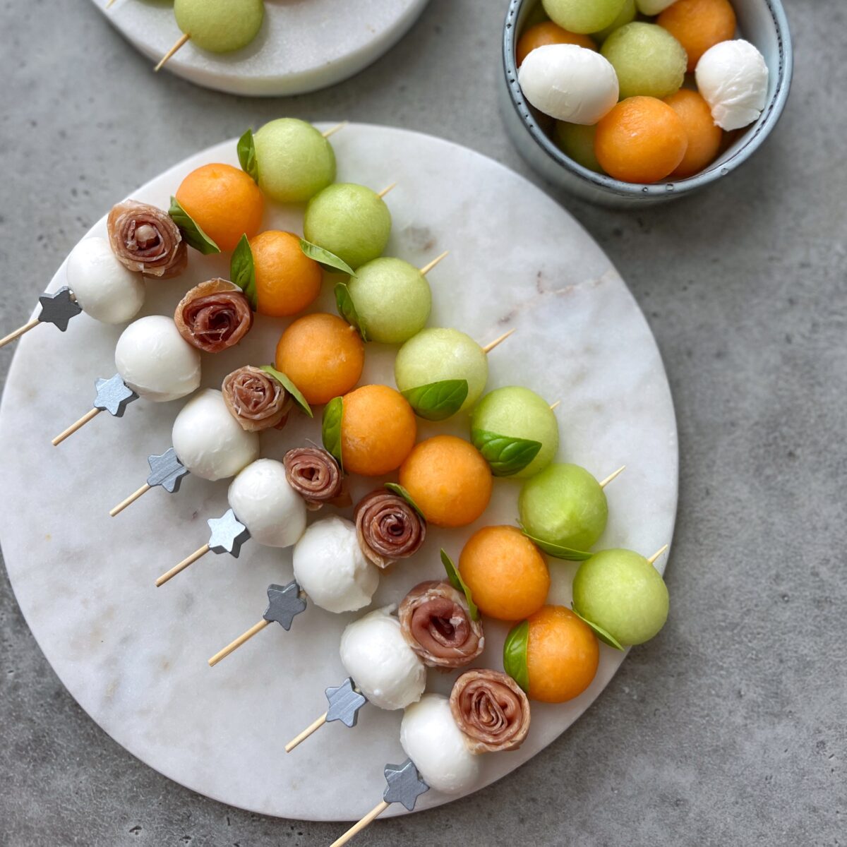 Wooden skewers with mozzarella balls, prosciutto, cantaloupe, and honeydew melon bites are garnished with fresh basil and arranged on a round marble platter.