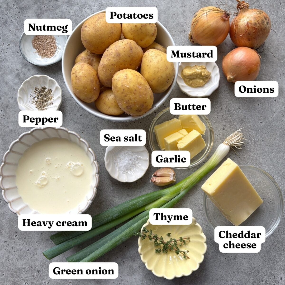 Ingredients for a delicious Potato and Onion Bake are arranged on a tabletop, including potatoes, onions, green onion, cheddar cheese, heavy cream, butter, garlic, mustard, thyme, nutmeg, pepper, and sea salt.