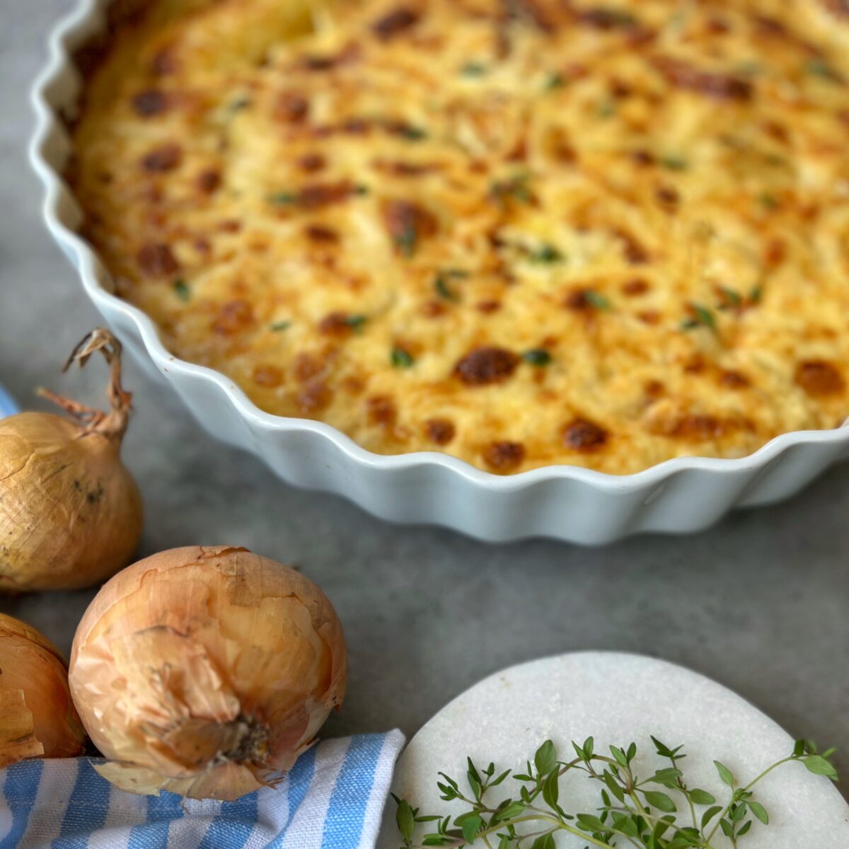 A baked gratin in a scalloped white dish sits on a gray surface. Beside it are three onions, fresh thyme sprigs on a round marble board, and a blue-and-white striped cloth.