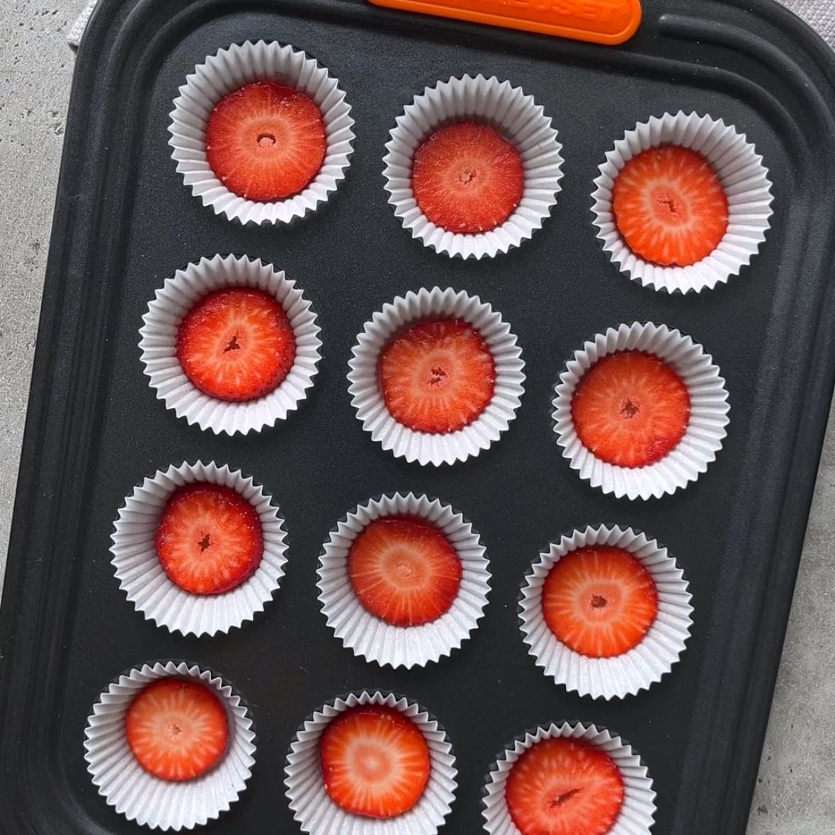 A muffin pan with twelve paper liners, each holding a single slice of strawberry, is shown from above on a gray surface.