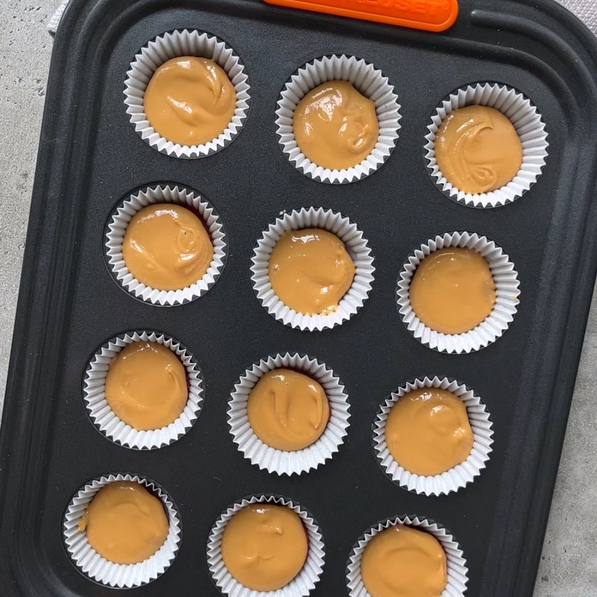 A muffin tin with 12 paper liners, each filled with peanut butter, sits on a gray surface.