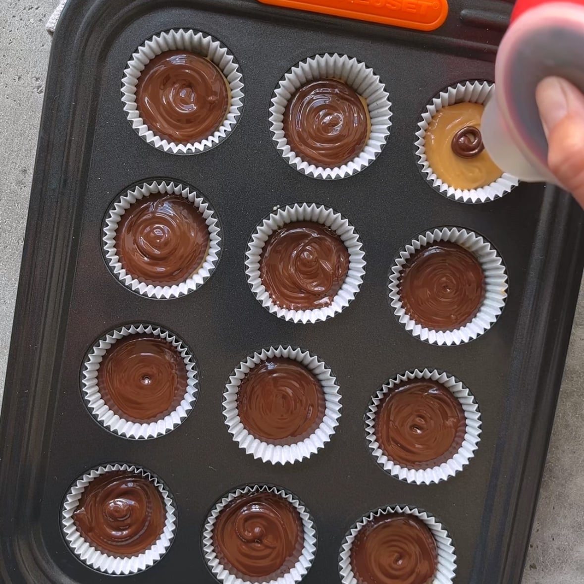 A muffin pan with paper liners, most filled with chocolate; a hand pours melted chocolate into the last top right liner.