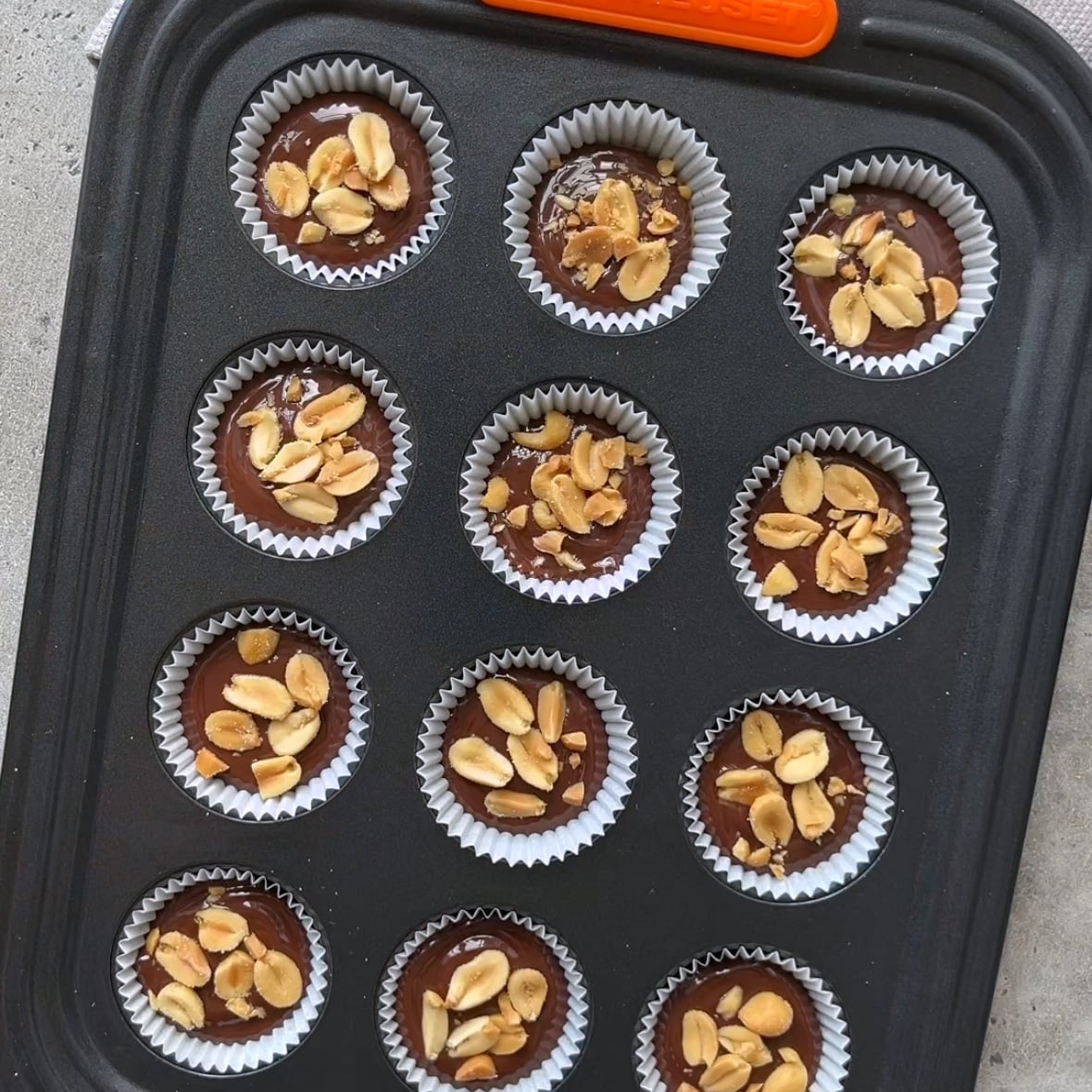 A muffin tin holds twelve chocolates in white liners, each topped with chopped peanuts, on a gray surface.
