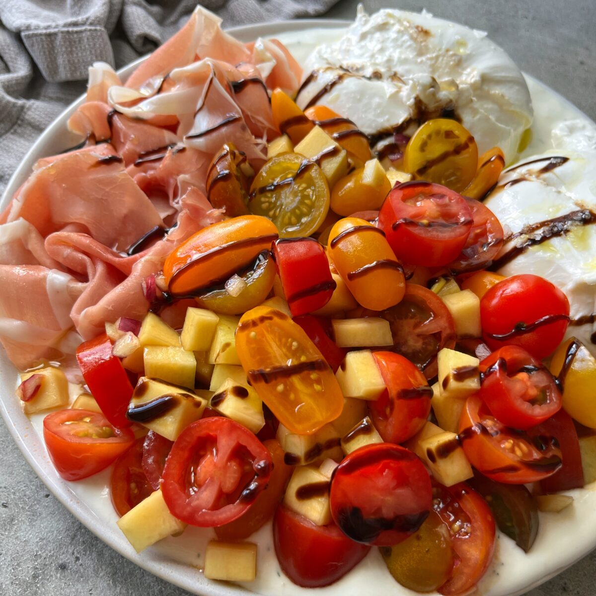 A plate with yogurt garlic dip topped with burrata cheese, prosciutto, cherry tomatoes, diced mango, and balsamic glaze.