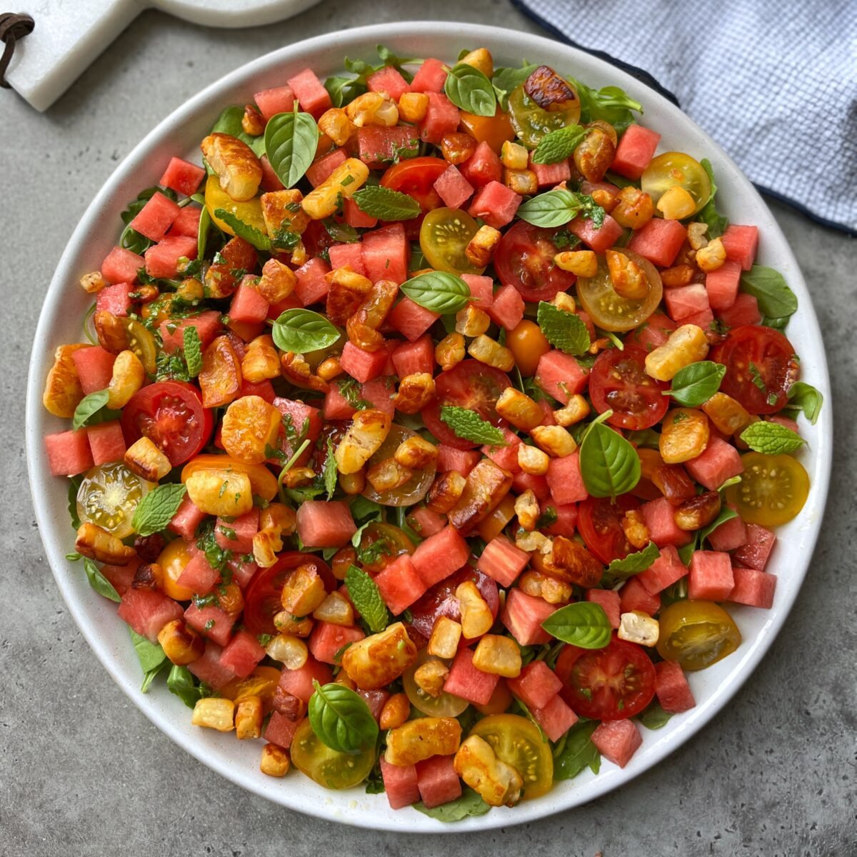 A plate of salad with cubed watermelon, halloumi cheese, cherry tomatoes, fresh basil, and mint leaves on a gray surface.