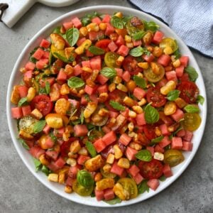 A plate of salad with cubed watermelon, halloumi cheese, cherry tomatoes, fresh basil, and mint leaves on a gray surface.
