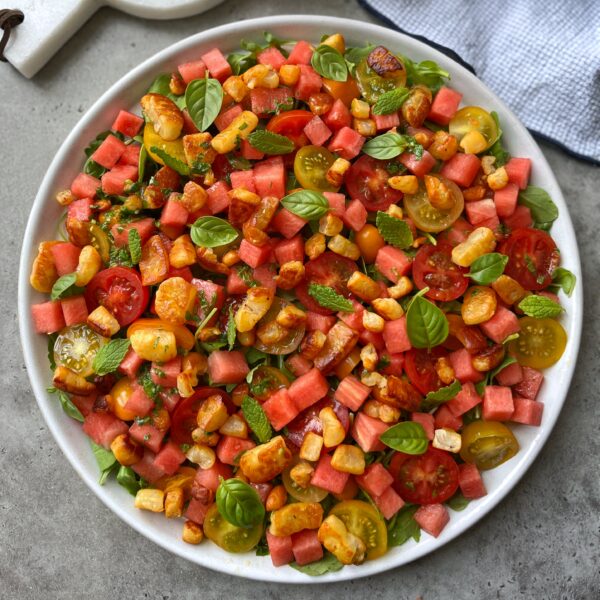 A plate of salad with cubed watermelon, halloumi cheese, cherry tomatoes, fresh basil, and mint leaves on a gray surface.