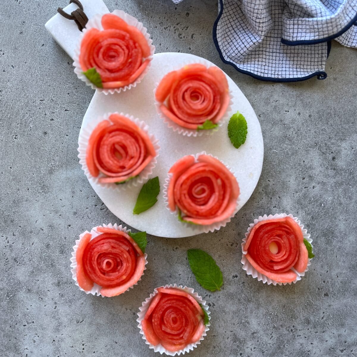 Seven rose-shaped watermelon bites arranged in white cupcake liners on a marble serving board, garnished with mint leaves.