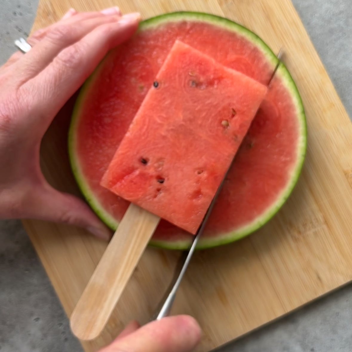 A hand slices a round watermelon with a knife while holding a rectangular watermelon popsicle on top.