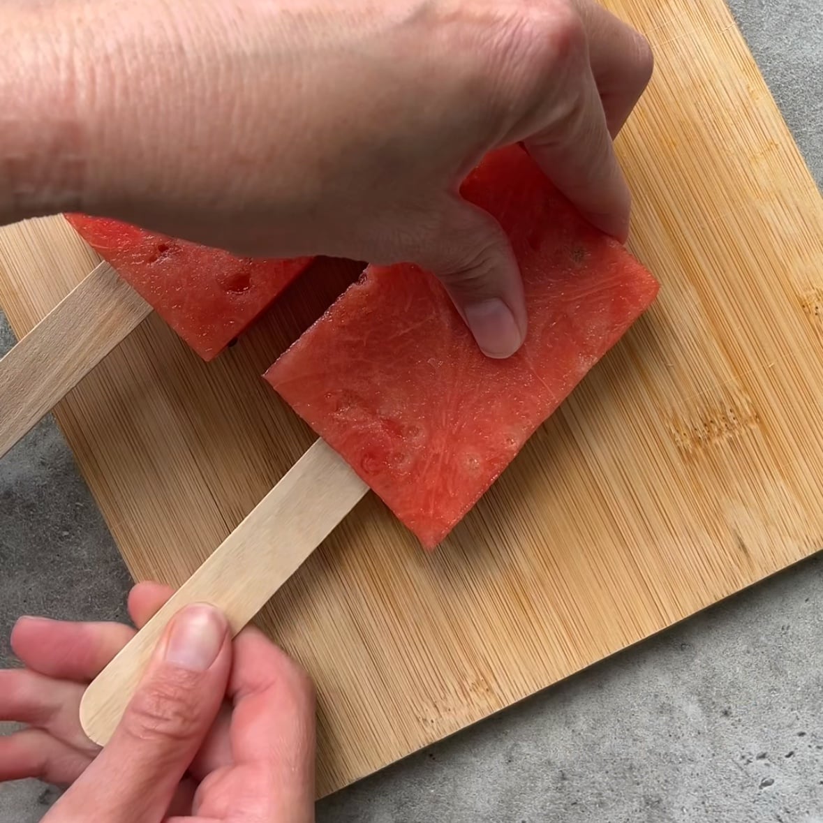 A hand holds a rectangular slice of watermelon on a wooden stick, placed on a wooden cutting board.