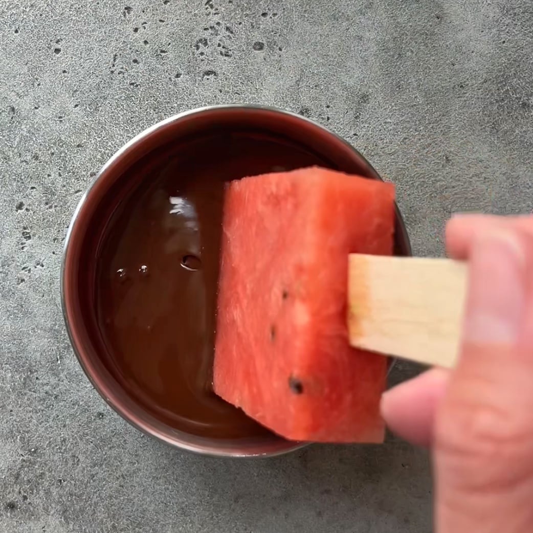 A hand holds one of the Watermelon Popsicles with Chocolate on a stick, dipping it into a bowl of rich chocolate sauce on a gray textured surface.