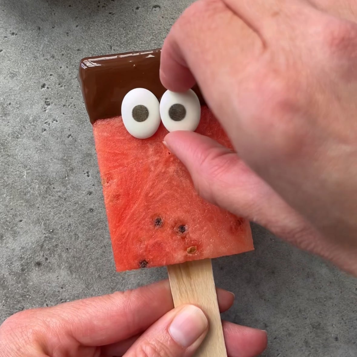 A hand places candy eyes on a Watermelon Popsicle with Chocolate, a watermelon slice dipped in chocolate and held on a wooden stick, over a gray surface.