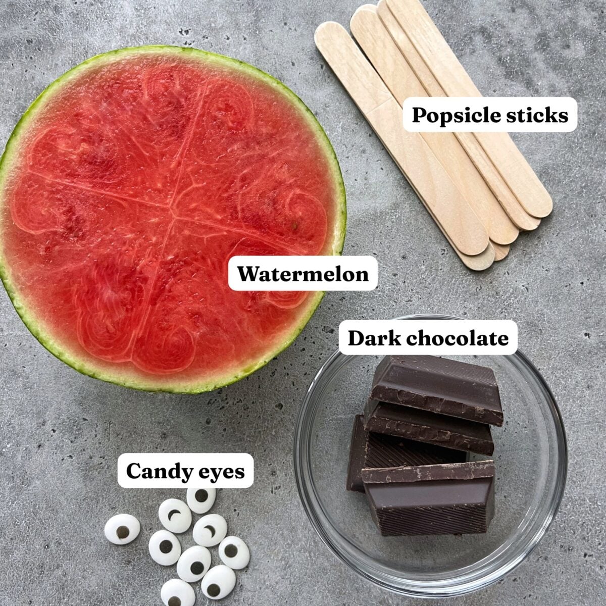 Half a watermelon, popsicle sticks, dark chocolate in a bowl, and candy eyes are arranged on a gray surface.