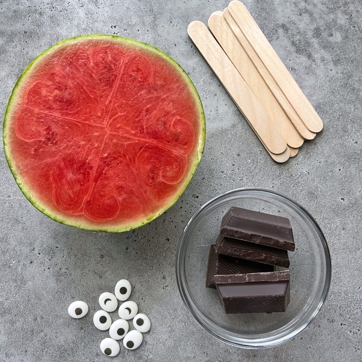 Half a watermelon, wooden popsicle sticks, a glass bowl with chocolate pieces, and candy eyes arranged on a gray surface.