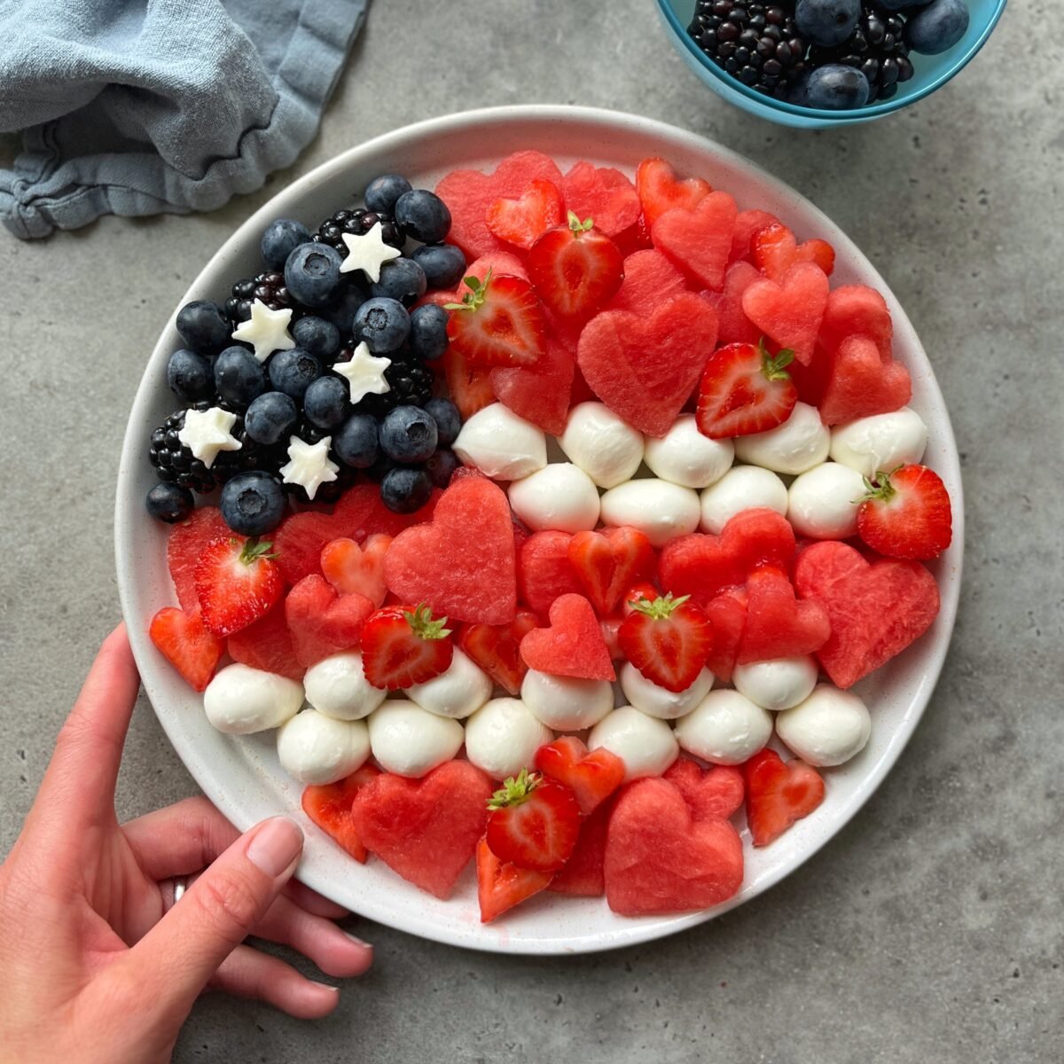 A plate arranged to resemble the American flag using blueberries, star-shaped cheese, mozzarella balls, heart-shaped watermelon, and strawberries. A hand holds the edge of the plate.