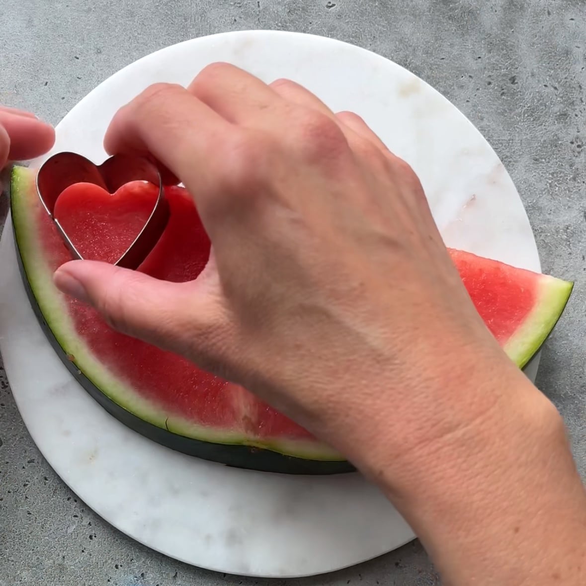 Hands cut out hearts from from a halved piece of watermelon.