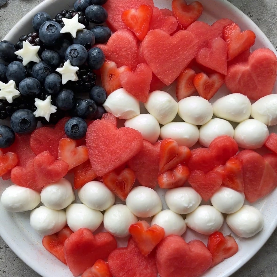 A 4th of July salad featuring blueberries and blackberries in the top left corner topped with mozzarella stars, strips of strawberries and watermelon, as well as strips of mozzarella balls arranged in the shape of an American flag.
