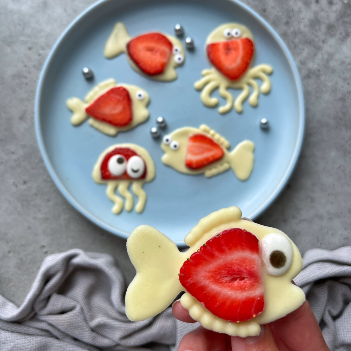 A hand holds a white chocolate fish-shaped treat with a strawberry slice and candy eye. On a blue plate in the background are more fun bites made from white chocolate, strawberries, and candy decorations—perfect for creative food recipes.