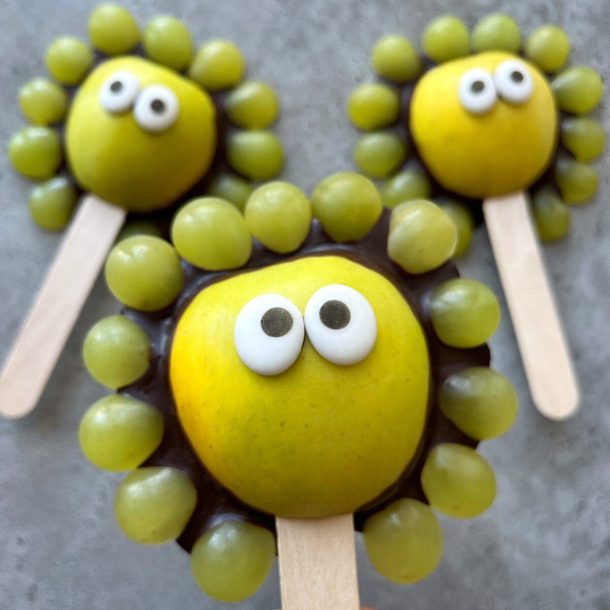 Three fruit snacks resembling cartoon faces, made from apple halves, grapes, candy eyes, and chocolate, each with a popsicle stick handle, arranged on a gray surface.