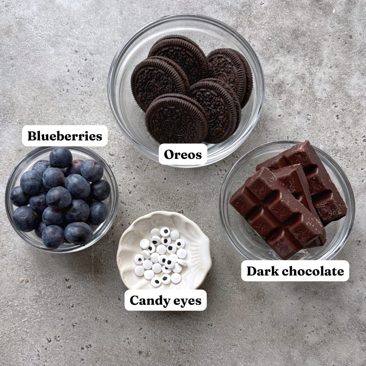 Four small bowls containing blueberries, Oreo cookies, dark chocolate pieces, and small candy eyes.