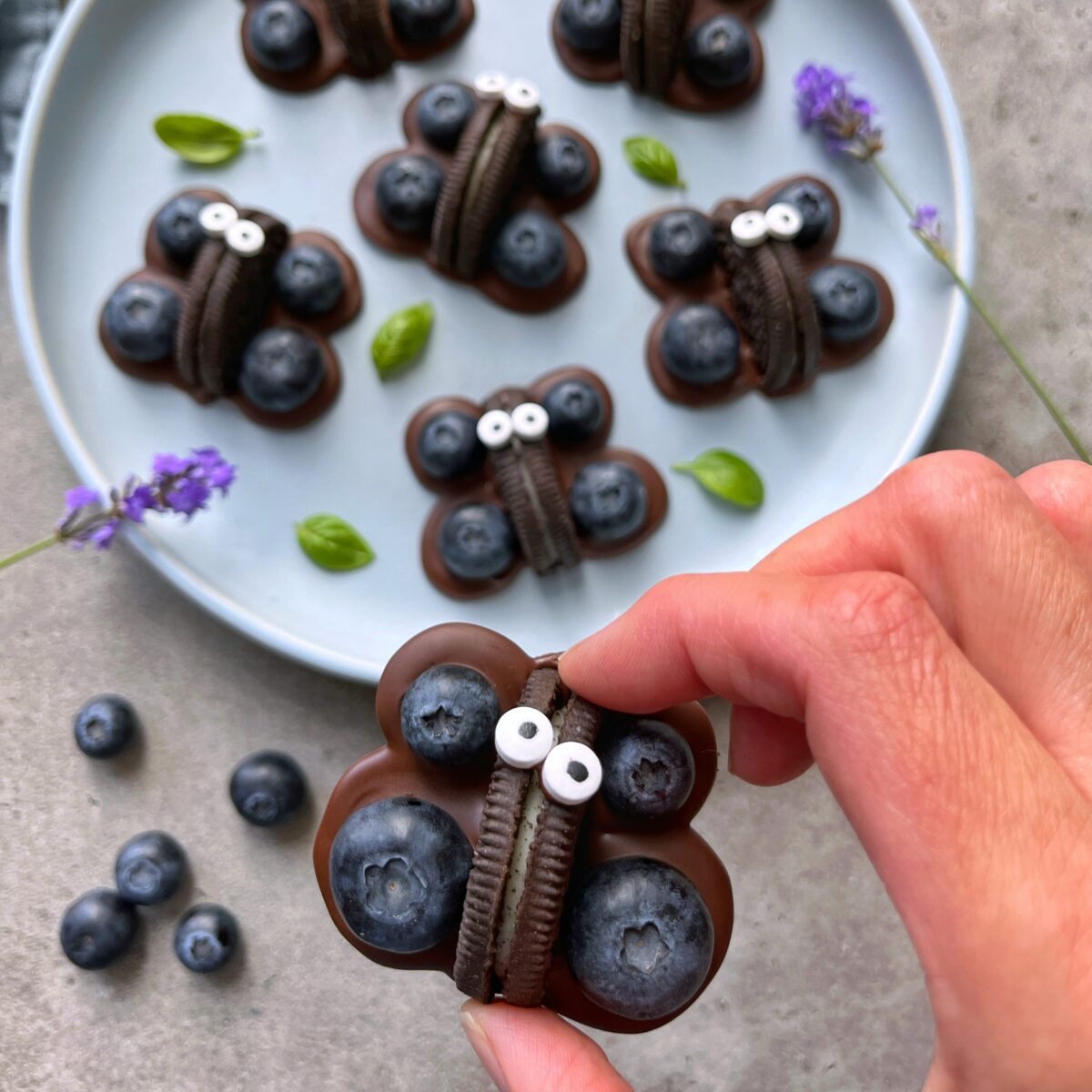 A hand holds a butterfly-shaped treat made with chocolate, blueberries, an Oreo cookie, and candy eyes. Similar bites rest on a plate in the background.