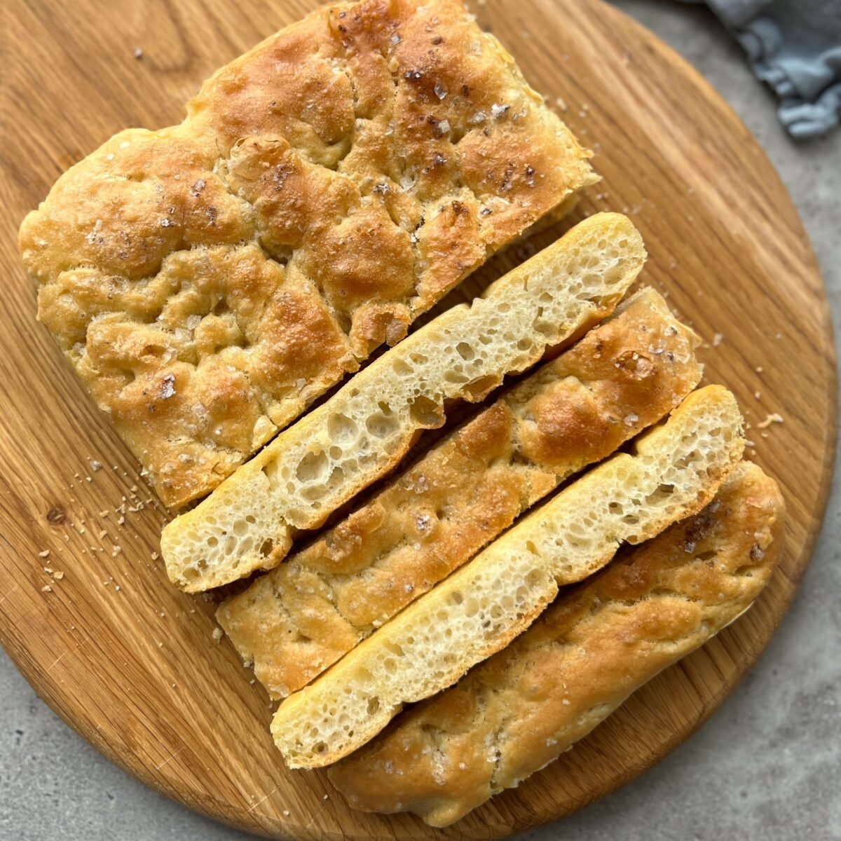A rectangular loaf of focaccia bread, featured in many food recipes, sits sliced on a round wooden board. The bread has a golden crust and a coarse, airy crumb.