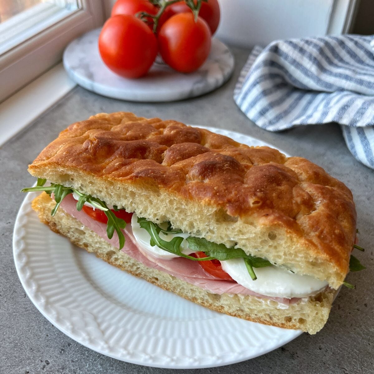 A sandwich with ham, mozzarella, tomato, and arugula on Easy No-Knead Focaccia sits on a white plate.