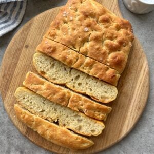 A sliced loaf of focaccia bread with a golden crust, displayed on a round wooden cutting board.