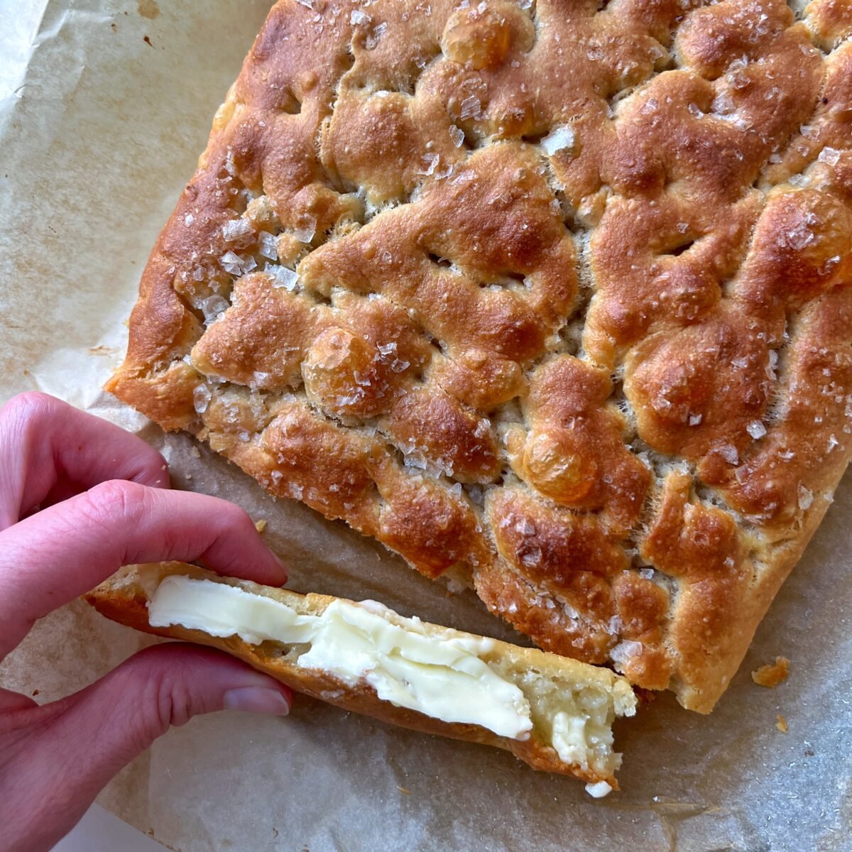 A hand holds a slice of focaccia bread with butter shown before a larger sea salt-topped loaf resting on parchment paper.