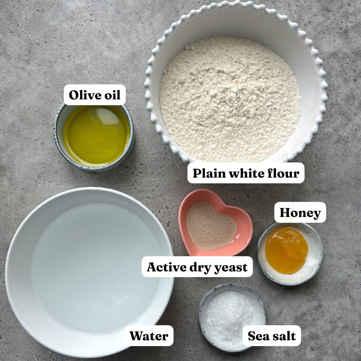 A bowl of flour, olive oil, water, yeast in a heart-shaped dish, honey, and sea salt, arranged on a gray surface.