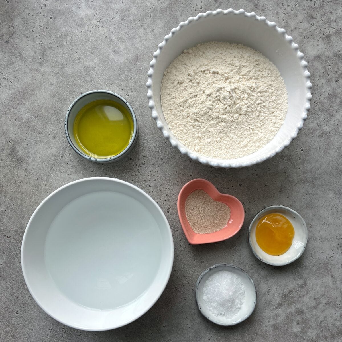 A bowl of flour, olive oil, water, yeast in a heart-shaped dish, honey, and sea salt, arranged on a gray surface.
