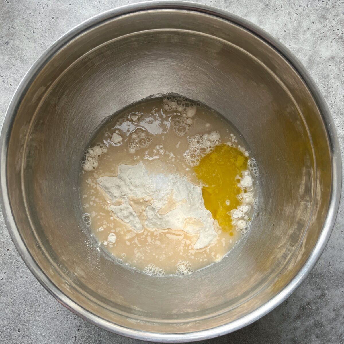 A metal mixing bowl containing flour, olive oil, sea salt, yeast, and water ingredients on a gray surface.