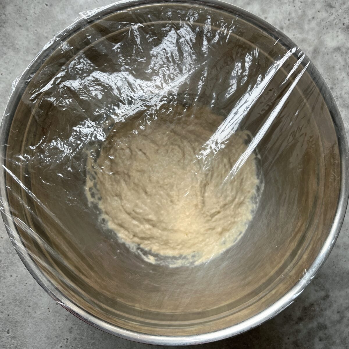 A metal bowl filled with focaccia dough is covered with plastic wrap, resting on a gray surface.