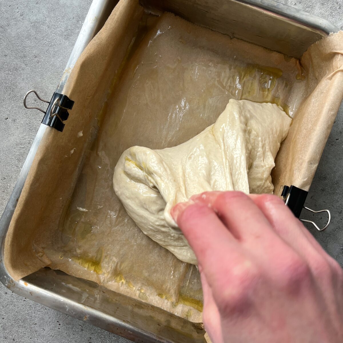 A hand stretches raw dough in a parchment-lined baking pan.