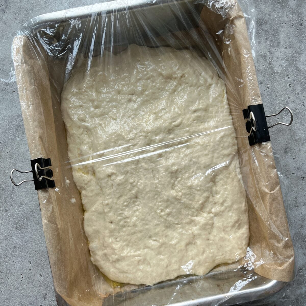A pan lined with parchment paper holds sticky focaccia dough, covered with plastic wrap and secured with binder clips, set on a gray surface.