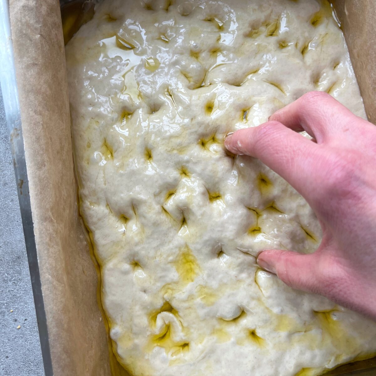 A hand presses dimples into Easy No-Knead Focaccia dough covered with oil in a parchment-lined baking pan.