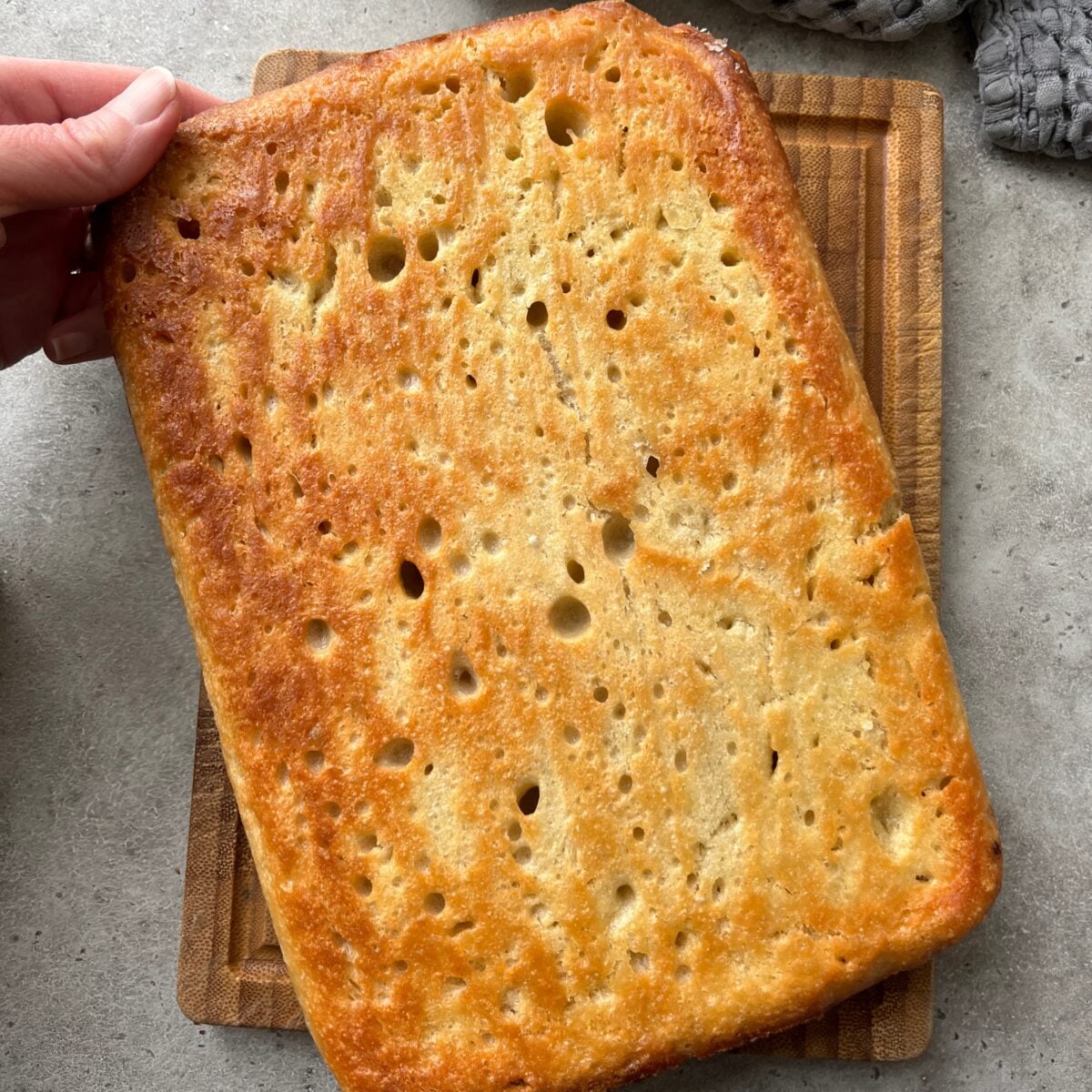 A rectangular, golden-brown Easy No-Knead Focaccia bread with visible holes sits on a wooden cutting board, held at one corner by a hand.