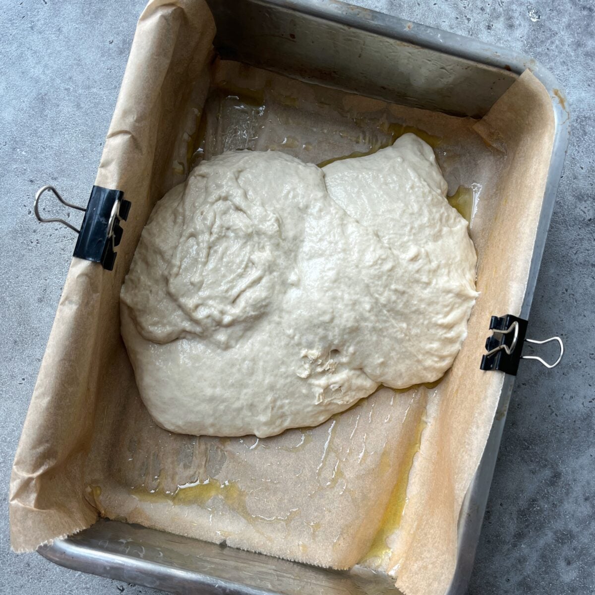 Focaccia dough is transferred to a rectangular baking pan, with parchment paper secured with binder clips. The dough sits atop a layer of olive oil.