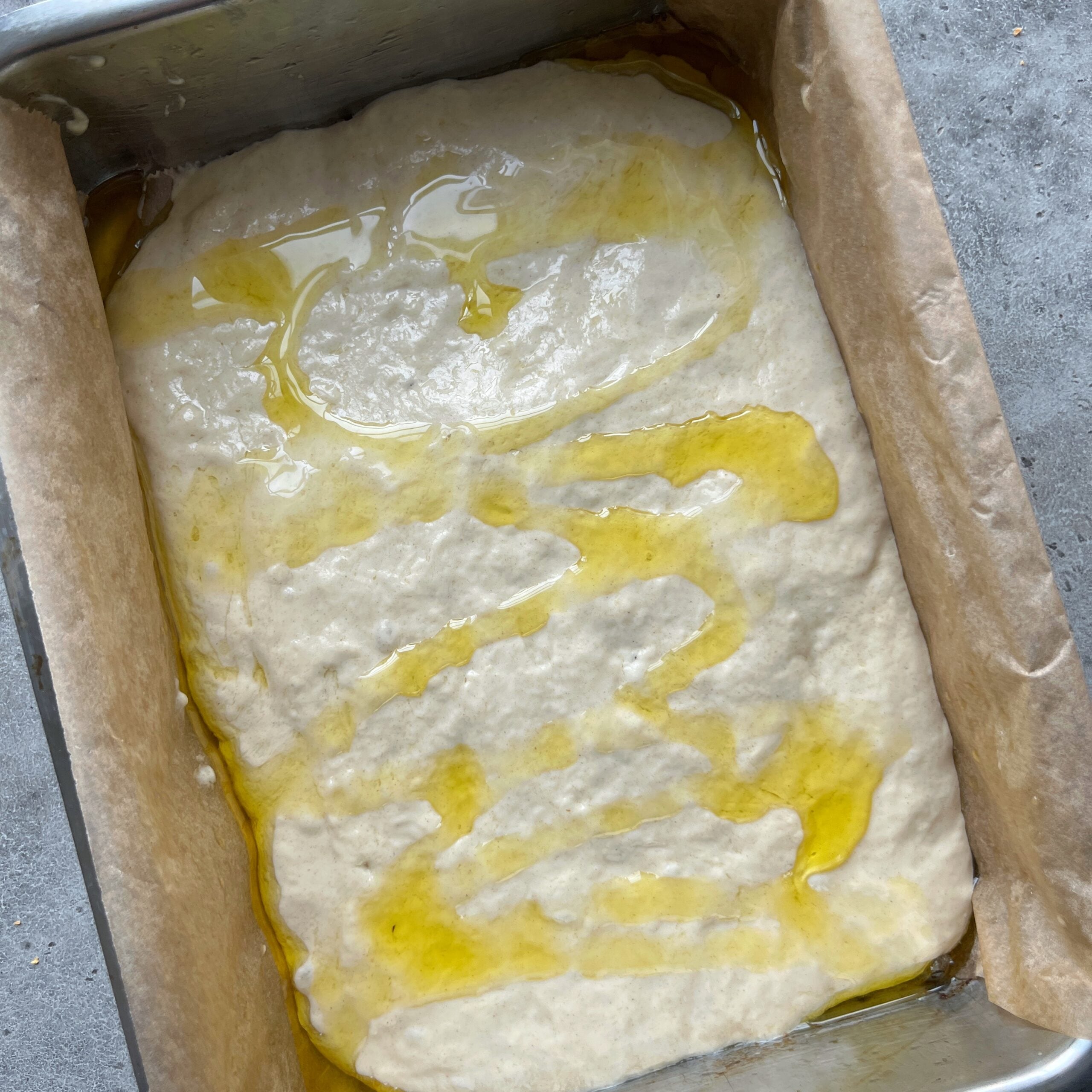 Easy No-Knead Focaccia dough rests in a parchment-lined pan, drizzled with olive oil ready to be dimpled.
