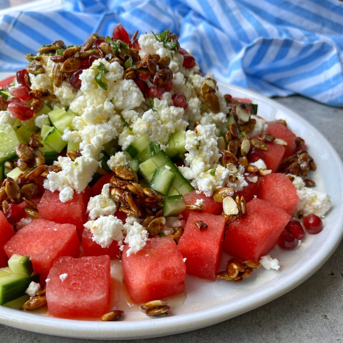 A plate of cubed watermelon, cucumber, crumbled feta cheese, pomegranate seeds, and caramelized pumpkin seeds with a blue and white striped cloth in the background.