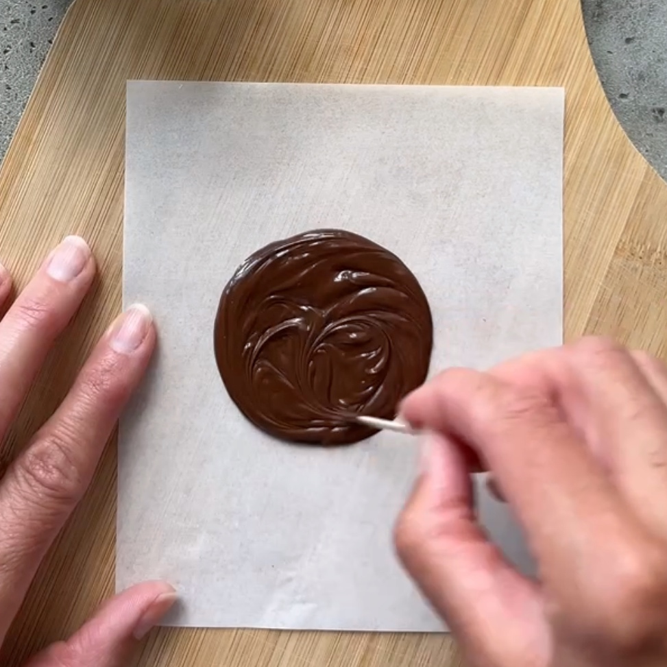 A toothpick spreads out melted dark chocolate into a perfect disc on parchment paper.