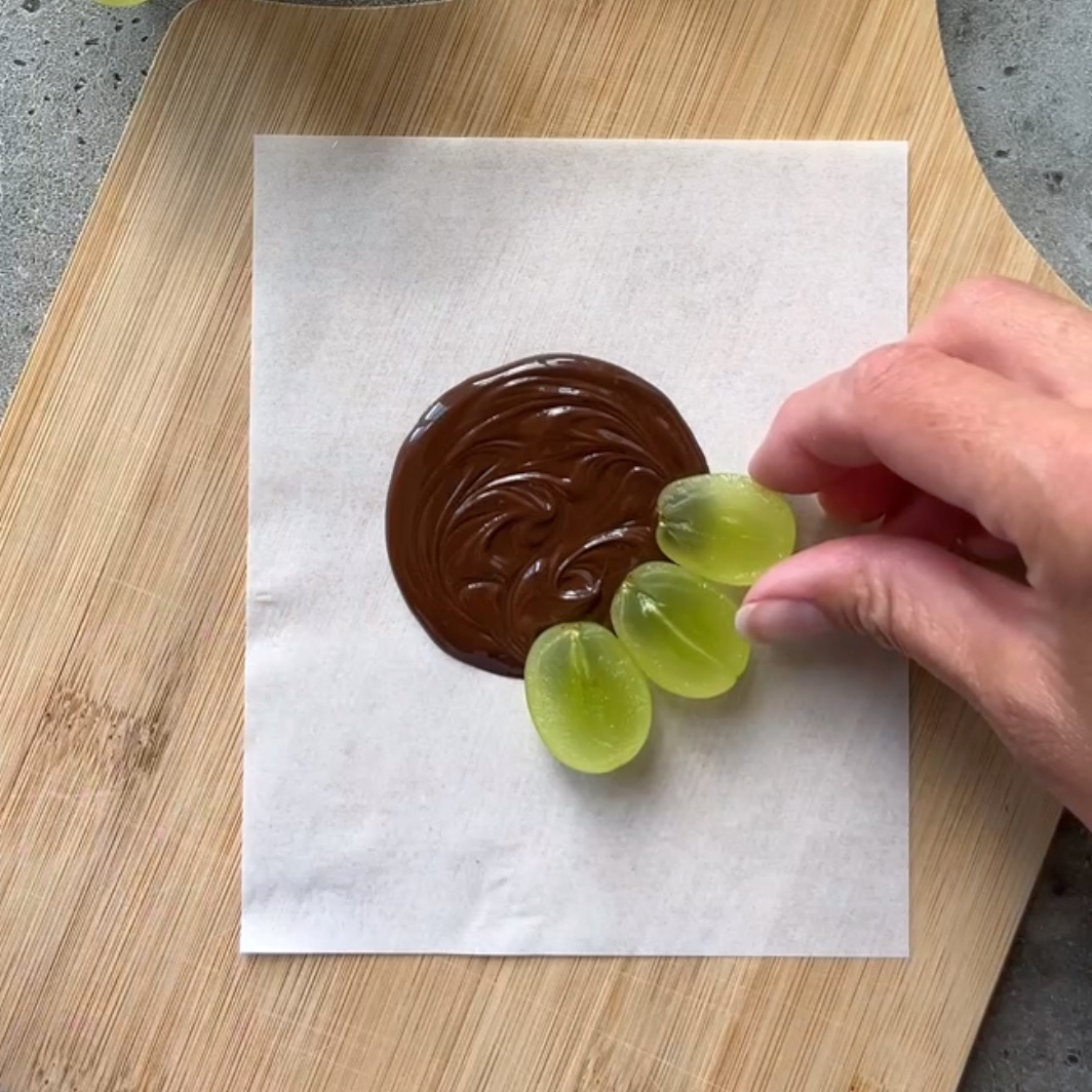 A hand arranges halved green grapes on the outer edge of a dark chocolate disc.