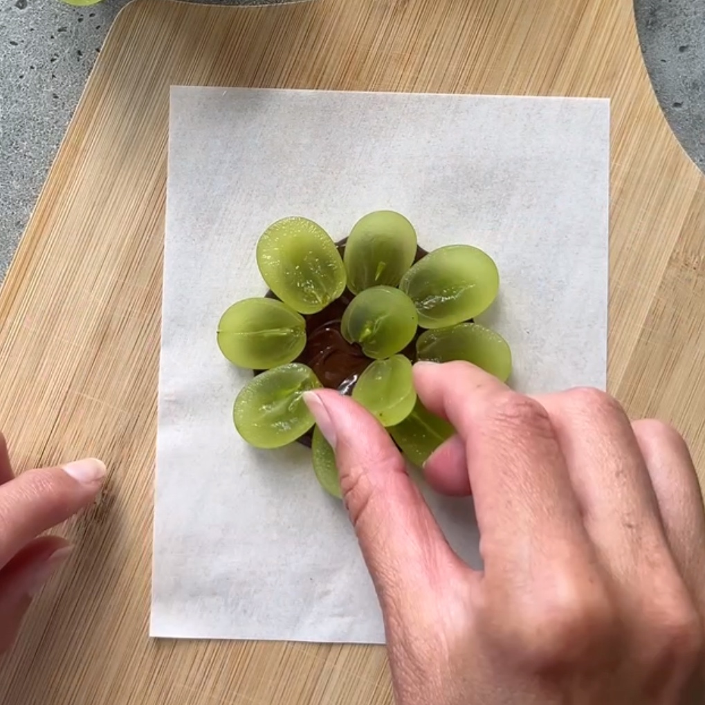 A hand places smaller grape halves inside the center of a dark chocolate disc lined with more halved grapes.
