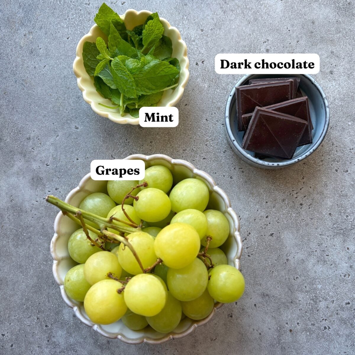Three bowls  containing fresh mint, dark chocolate squares, and green grapes.
