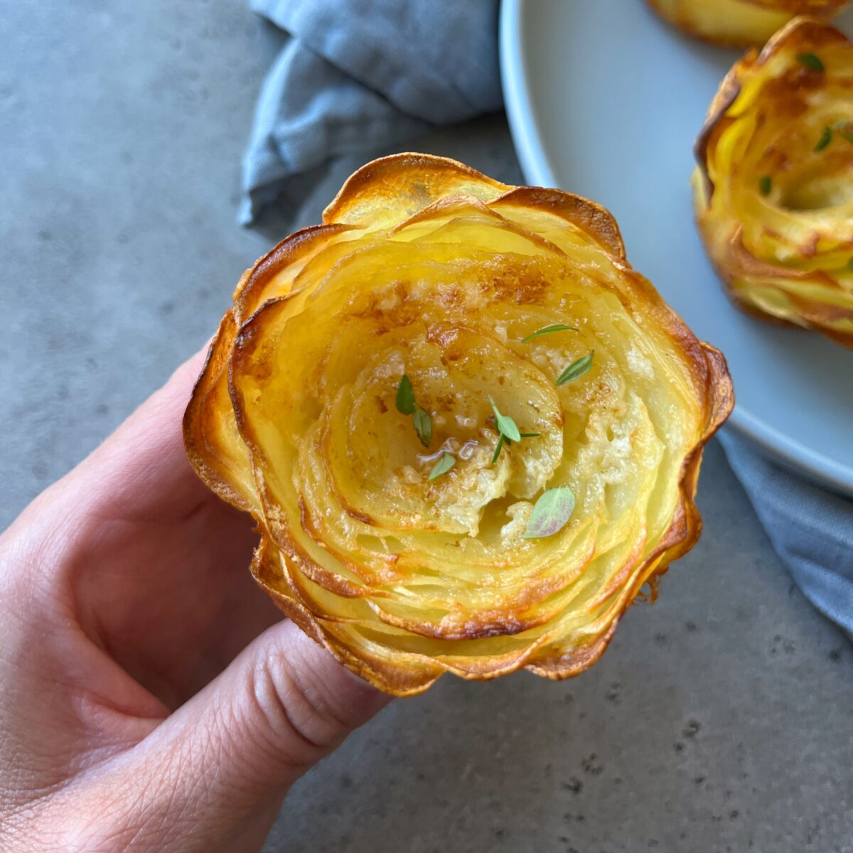A closeup of a crispy and golden potato rose, garnished with fresh thyme. There are more potato roses and a blue tea towel nearby.