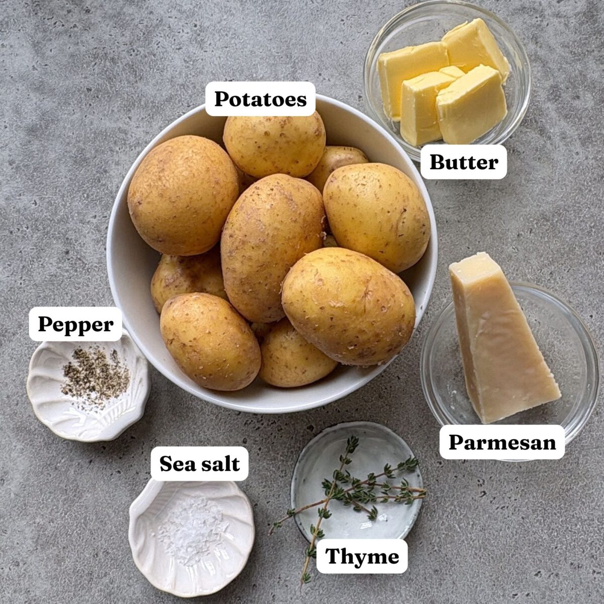 A bowl of whole potatoes surrounded by labeled ingredients including butter, parmesan cheese, thyme, sea salt, and ground pepper on a gray surface.